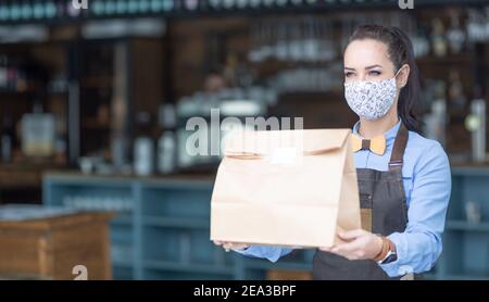 Restaurantmitarbeiter übergibt während der Pandemie von Covid-19 Lebensmittel, die in einer Papiertüte mit Gesichtsmaske verpackt sind. Stockfoto