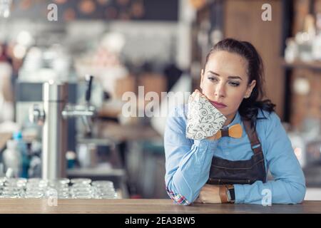 Traurige Geschäftsbesitzerin hält Gesichtsmaske in der Hand und lehnt sich an die Bar in ihrem leeren Café. Stockfoto