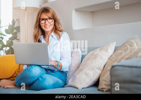 Aufnahme einer Frau mittleren Alters, die zu Hause einen Laptop auf dem Sofa benutzt. Geschäftsfrau, die von zu Hause aus arbeitet. Stockfoto
