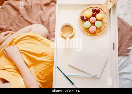 Blick von oben auf das Frühstück im Bett. Mädchen in gelbem Kleid sitzt auf dem Bett mit Tablett mit Kaffee, Dessert und leeres Notizbuch. Weißes Papier und Bleistift zum Schreiben. Makronen und Kirschen auf einem Holzbrett. Stockfoto