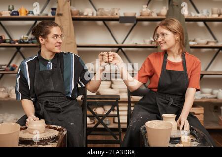 Vorderansicht Porträt von zwei jungen Handwerkern stoßen Fäuste während der Herstellung von Keramik in Töpferwerkstatt mit Töpferscheibe, kopieren Raum Stockfoto