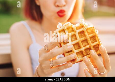 Eine glückliche Frau in einem Stadtpark ist beim Mittagessen und hält ein Wiener Waffelbrot. Moderne leckere und kalorienreiche Street Food Stockfoto