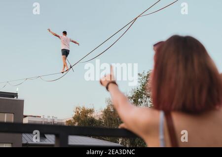 Männlicher Athlet demonstriert seine Fähigkeiten, indem er auf einer schmalen Slackline balanciert, die zwischen zwei Gebäuden in der Stadt gespannt ist. Das Publikum beobachtet die Tricks w Stockfoto