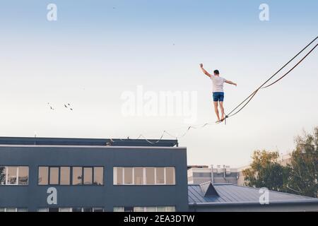 Männlicher Athlet demonstriert seine Fähigkeiten, indem er auf einer schmalen Slackline balanciert, die zwischen zwei Gebäuden in der Stadt gespannt ist. Das Publikum beobachtet die Tricks w Stockfoto