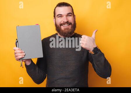 Foto von gutaussehenden fröhlichen Mann zeigt Daumen nach oben und hält Planer. Stockfoto