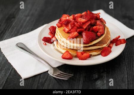 Ein Stapel von drei Pfannkuchen auf einem Teller, gekrönt mit geschnittenen Erdbeeren, mit einer Gabel und Serviette auf einem dunklen Holzhintergrund. Stockfoto