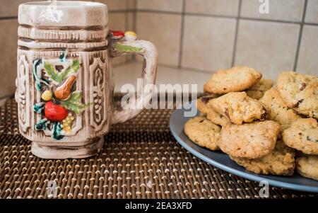 Ein grauer Teller mit Schokoladenstückchen Plätzchen und einem bunten Becher daneben auf Weidenoberfläche Stockfoto