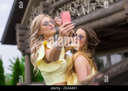 Porträt von zwei hübschen jungen Brautjungfern in gelben Kleidern in der Nähe von einem modischen Holzhaus. Menschen Lifestyle-Konzept. Machen Selfie auf Handy p Stockfoto