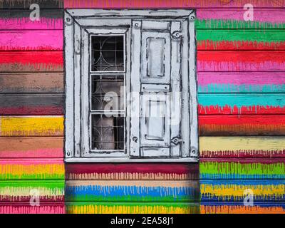 Vintage-Fenster, mit weißen und schwarzen Farben bemalt, in der bunten Wand geschält alten Holzhaus. Fragment der alten, mehrfarbigen, strukturierten Wand. Stockfoto