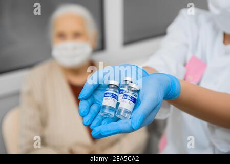 Fläschchen mit Covid-Impfstoff in den Händen eines Arztes Vor dem Hintergrund einer älteren Frau Stockfoto