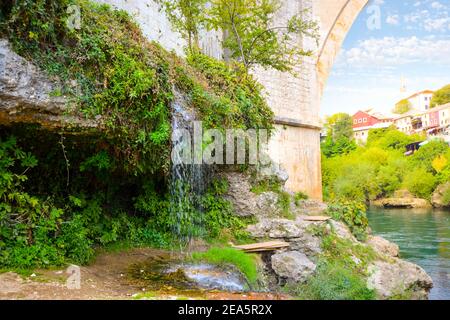 Ein kleiner Wasserfall neben der historischen Old Mostar Brücke oder Stari Most mit dem Dorf Mostar, Bosnien-Herzegowina mit Blick über den Fluss Neretva Stockfoto