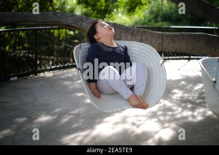 Ein süßes junges asiatisches Mädchen, das in einer Schaukel unter einem großen Baum in einem Garten sitzt, müde ist und nach einem langen Spieltag einschläft. Stockfoto