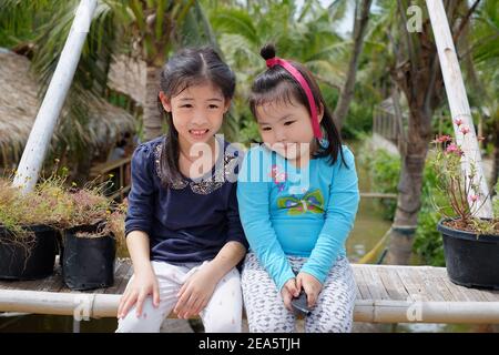 Portrait von zwei niedlichen jungen asiatischen Mädchen, die Schwestern sind, sitzen zusammen in einer Bank im Freien, genießen ihre Zeit zusammen, lächelnd. Stockfoto