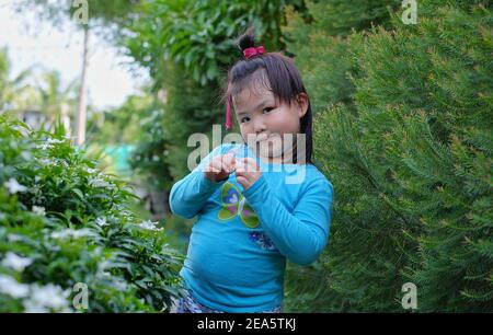 Ein süßes junges asiatisches Mädchen posiert für die Kamera in einem Garten mit Bäumen im Hintergrund, lächelnd, aber schüchtern. Stockfoto