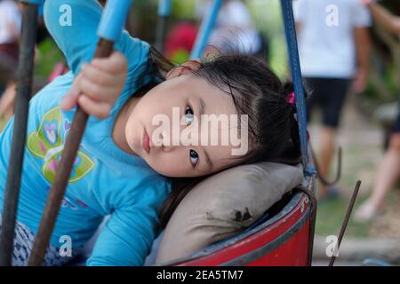 Ein süßes junges asiatisches Mädchen, das im Rücken eines Dreirads sitzt, ihren Kopf auf dem Kissen des Rücksitzes ruht und sich entspannt und schläfrig fühlt. Stockfoto