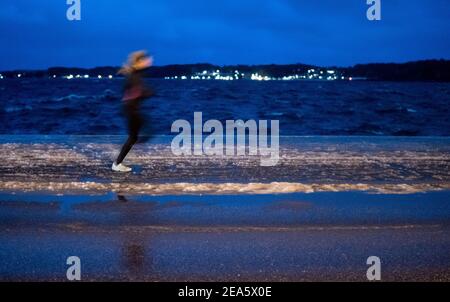 Kiel, Deutschland. Februar 2021, 08th. Ein Jogger am frühen Morgen läuft nahe dem Ufer der Ostsee auf einer Straße, auf der sich eine Eisschicht gebildet hat, nachdem starke Winde das Wasser der Ostsee auf die Straße geweht haben. Quelle: Axel Heimken/dpa/Alamy Live News Stockfoto