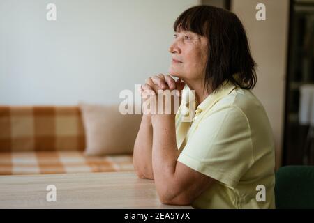 Eine reife Frau sitzt an einem Tisch in der Küche Am Fenster mit nachdenklicher Optik Stockfoto