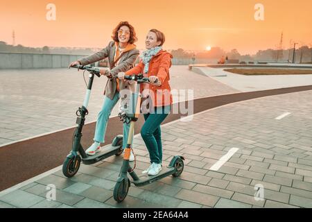 Zwei Freundinnen fahren moderne gemietete Elektroroller durch die Straßen der Stadt. Transport und Lifestyle und Freundschaftskonzept Stockfoto