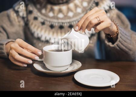 Eine Frau in einem Winterpullover gießt laktosefreie Milch oder Sahne in ein heißes Getränk - Kaffee oder Tee. Konzept eines gemütlichen Cafés und der Gehalt an tierischen Fetten i Stockfoto