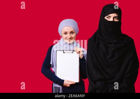 Muslimische Mädchen auf rotem Hintergrund. Ein junges Mädchen hält einen Ordner, weißes Blatt, lächelt und schaut auf die Kamera, ein anderes verdächtig aussehend schwarz Stockfoto