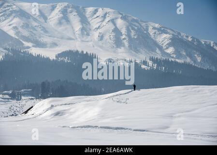 Ein Mann geht entlang einer schneebedeckten Wiese in Gulmarg. Gulmarg ist eine Stadt, eine Bergstation, ein beliebtes Skiziel und ein mitteiltes Gebietskomitee im Baramulla Distrikt Jammu und Kaschmir. Die Stadt liegt in der Pir Panjal Range im westlichen Himalaya und liegt innerhalb der Grenzen des Gulmarg Wildlife Sanctuary. Stockfoto