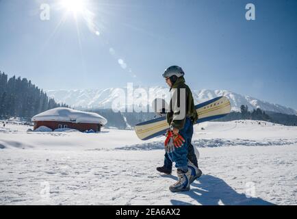 Ein Snowboarder wandert entlang einer schneebedeckten Wiese in Gulmarg. Gulmarg ist eine Stadt, eine Bergstation, ein beliebtes Skiziel und ein mitteiltes Gebietskomitee im Baramulla Distrikt Jammu und Kaschmir. Die Stadt liegt in der Pir Panjal Range im westlichen Himalaya und liegt innerhalb der Grenzen des Gulmarg Wildlife Sanctuary. Stockfoto