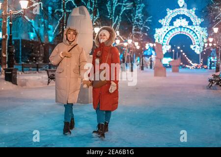 Glückliche Freundinnen gehen im abendlichen Winterpark auf dem Bürgersteig vor dem Hintergrund der Weihnachts- und Neujahrsdekorationen in Ufa, Russland Stockfoto