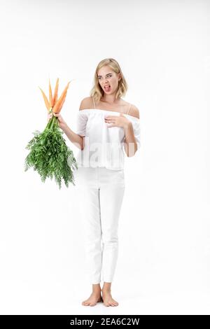 Blonde Frau in einer weißen Bluse hält eine Karotte mit grünen Blättern auf weißem Hintergrund. Mädchen fühlt sich schlecht von Karotten und Diäten Stockfoto