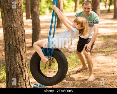 Vater schiebt Kind Tochter auf Reifenschaukel im Sommerwald. Aktives Familienwochenende. Hochwertige Fotos Stockfoto