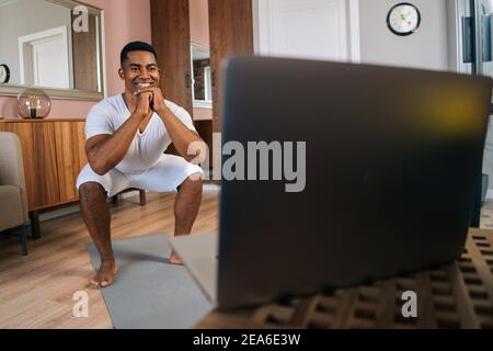 Frontansicht der fröhlichen afroamerikanischen Mann Ausübung und tun Kniebeugen online vor dem Laptop. Stockfoto