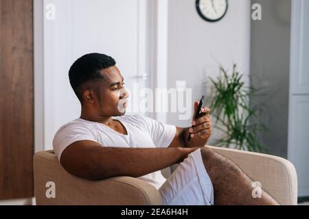 Seitenansicht eines entspannten afroamerikanischen Mannes, der auf einem bequemen Sessel sitzt und ein Mobiltelefon benutzt. Stockfoto