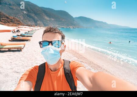 Ein männlicher Tourist, der eine Sonnenbrille und eine schützende medizinische Maske trägt Spaziergänge und Ruhepausen an einem Sandstrand am Mittelmeer Meer während eines Coronavirus pandemi Stockfoto