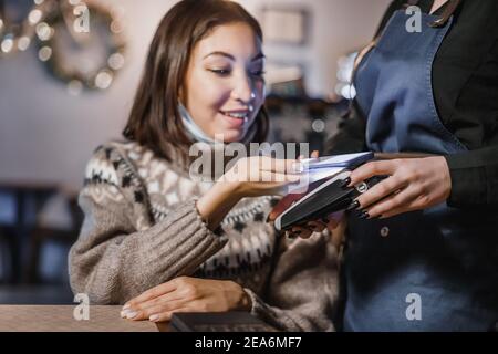 Eine Frau zahlt für das Abendessen in einem Café mit NFC-Technologie in ihrem Smartphone, indem sie es auf dem Barista POS-Terminal befestigt Stockfoto