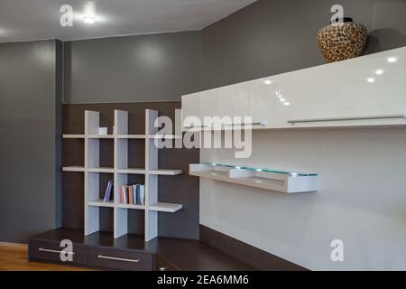 Modernes Interieur des Wohnzimmers in Luxus-Wohnung. Regal aus dunklem Holz mit Schubladen. Bücherregal mit Büchern. Weiße Schränke. Stockfoto