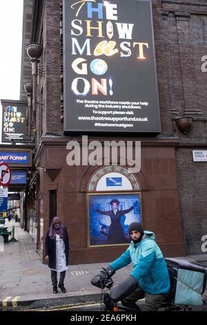 Deliveroo Kurier stoppt neben der Show Muct Go on Schild vor dem geschlossenen Prince Edward Theater in Soho, das Mary Poppins im West End zeigt, während die nationale Coronavirus-Sperre drei weiter geht und die Theater am 28th. Januar 2021 in London, Großbritannien geschlossen bleiben müssen. Nach der Zunahme der Fälle im Winter, einschließlich einer neuen britischen Variante von Covid-19, rät diese landesweite Sperre allen Bürgern, der Botschaft zu folgen, zu Hause zu bleiben, den NHS zu schützen und Leben zu retten. Stockfoto