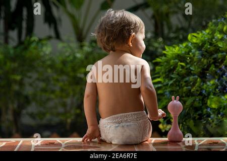 Ein charmantes Kleinkind sitzt mit einem Spielzeug auf den Stufen eines Hauses im Sonnenlicht. Rückansicht. Stockfoto