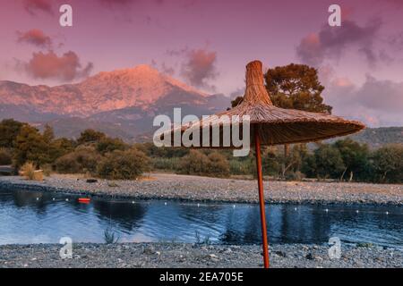 Sonnenuntergang in rosa Schatten mit Sonnenliege und Sonnenschirm am Strand und Berg Tahtali im Hintergrund. Tourismus und Erholung im Ferienort Tekirova an Stockfoto