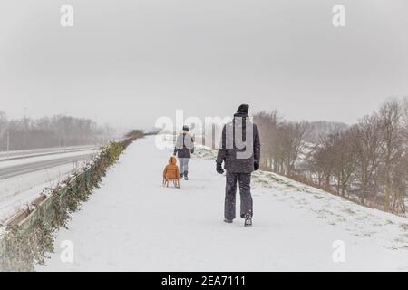 Familie mit Kind auf einem Schlitten Wandern im Schnee auf einem Deich in Ede, Gelderland in den Niederlanden Stockfoto