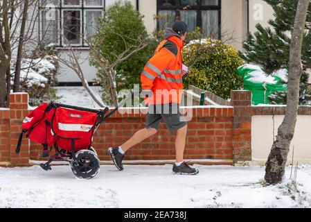 Southend on Sea, Essex, Großbritannien. Februar 2021, 8th. Der Schnee von Storm Darcy ist gefroren und hat die Bedingungen tückisch gemacht. Postarbeiter setzen ihre Runden im Schnee fort und liefern Post, einige in Shorts. Männlicher Postbote, der einen Wagen in einem Wohngebiet zieht Stockfoto