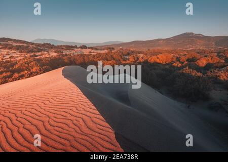 Majestätische Aussicht auf orangefarbene Sanddünen und Hügel glüht in den Strahlen des warmen Sonnenuntergangs in der Nähe von Patara Strand in der Türkei. Büsche und entfernte Berge im Bac Stockfoto