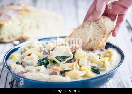 Frische Tortellini Suppe mit italienischer Wurst, Spinat und Karotten. Von Hand eintauchen hausgemachtes hausgemachtes Brot. Selektiver Fokus auf Pasta mit verschwommenem Hintergrund Stockfoto