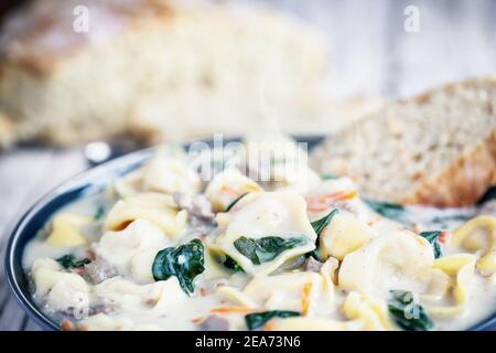 Frische Tortellini-Suppe mit italienischer Wurst, Spinat und Karotten. Serviert mit hausgemachtem hausgemachtem Brot auf einem weißen Holztisch. Selektiver Fokus. Stockfoto