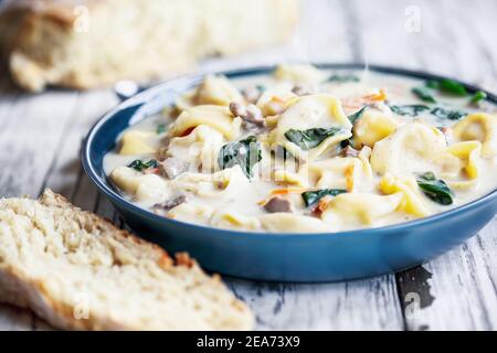 Frische Tortellini-Suppe mit italienischer Wurst, Spinat und Karotten. Serviert mit hausgemachtem hausgemachtem Brot auf einem weißen Holztisch. Selektiver Fokus. Stockfoto