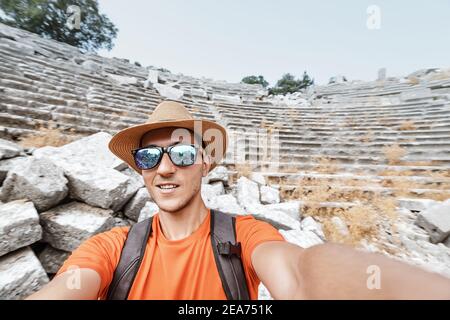 Ein männlicher Reisender mit Sonnenbrille und Hut nimmt ein Selfie gegen die Ruinen eines antiken Amphitheaters im Termessos Nationalpark in der Türkei. Stockfoto