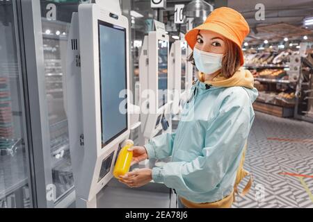 Eine Frau in einer medizinischen Schutzmaske kauft eine Flasche frischen Saft an einer Selbstbedienungskasse in einem Supermarkt. Das Konzept der Coronavirus-Ökonomie Stockfoto