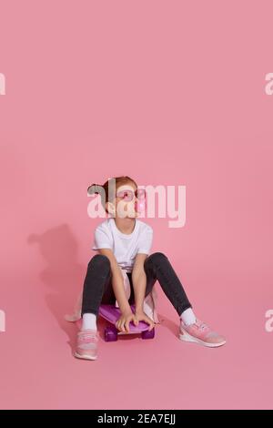 Cool Rotschopf kleines Kind Mädchen mit Kaugummi und rosa Sonnenbrillen auf Skateboard sitzen Stockfoto