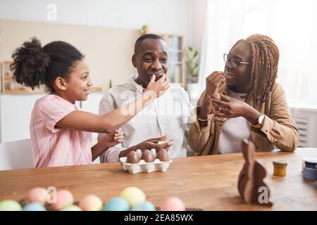 Portrait von liebevollen afroamerikanischen Familie Herstellung Schokolade Ostern Dekorationen und Verkostung sie beim Sitzen an Holztisch in gemütlichen Hause Innen Stockfoto