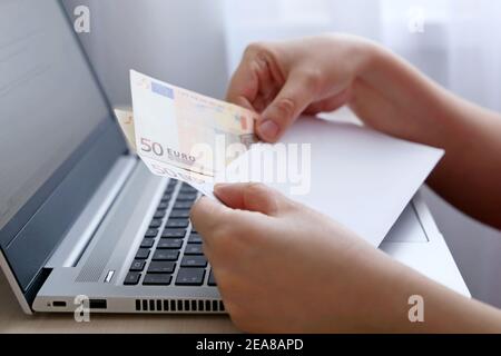 Briefumschlag mit Euro-Banknoten in weiblichen Händen. Frau zieht Geld aus einem Umschlag auf Laptop-Hintergrund, Löhne, Bonus oder Bestechung Konzept Stockfoto