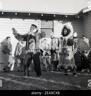 1960, historisch, an der Außengrenze von Anchorage, Alaska, USA, außerhalb eines einstöckigen Holzgebäudes, Ureinwohner in traditioneller Kleidung aus Tierfellen und Pelzen, die einen Tanz aufführen. Alaska wurde 49th der Staat der USA im Jahr 1959, aber es wurde von verschiedenen indigenen Völkern für Tausende von Jahren vor der Ankunft der Europäer besetzt. Da der Staat eine Seegrenze zu Russland hat, waren es sie, die sich als erste niederließen, was als russisches Amerika bekannt wurde. Die indigenen Völker machen über 15% der Bevölkerung aus und die alaskischen Ureinwohner spielen eine wichtige Rolle in der lokalen Regierung Stockfoto