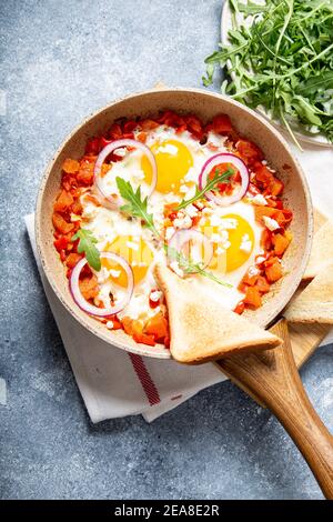 Leckere und gesunde Shakshuka in einer Bratpfanne. Eier pochiert in würziger Tomatensauce. Spiegeleier mit Toast, Rucola, lila Zwiebel und Käse Stockfoto